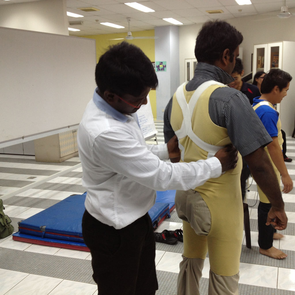 A clinician adjusts the back of a gold-colored TheraTogs garment on another participant during a hands-on training session. The setting includes mats and other participants practicing garment fitting techniques for postural and motor control.