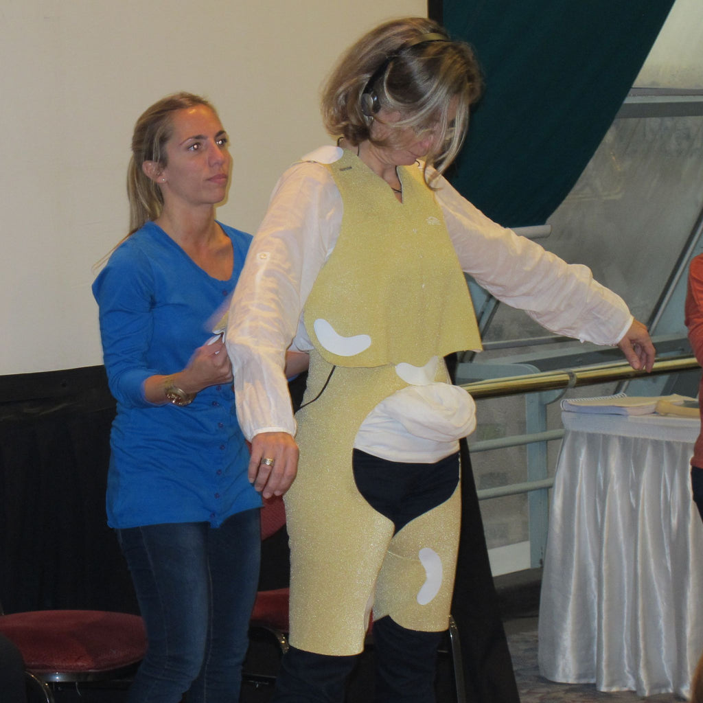 Two clinicians in a training session, with one clinician assisting a participant wearing a gold-colored TheraTogs garment. The participant stands with arms extended, demonstrating the fitting process of the therapeutic garment for postural alignment.